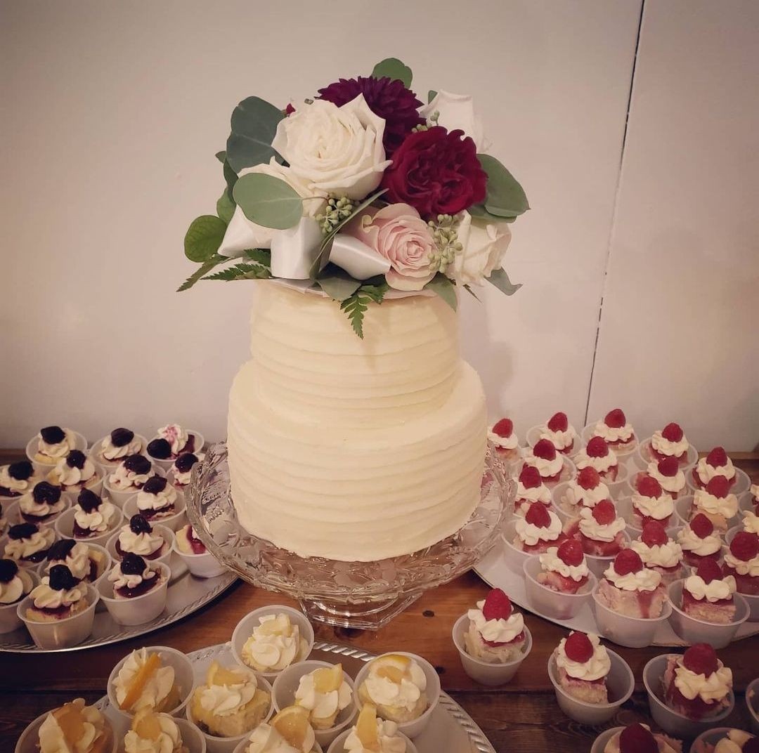 Two-tiered white cake topped with flowers, surrounded by various small cupcakes with different toppings.